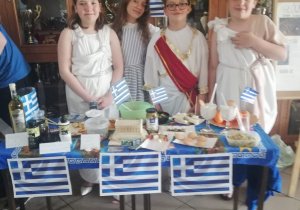 Na pierwszym planie stoi stolik przykryty niebieskim obrusem. Na stoliku stoją potrawy związane z Grecją. Za stolikiem stoja uczniowie przebrani za greckich bogów. W tle szafa z pucharami.